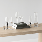 A set of assorted clear glassware, featuring the Orrefors Metropol Champagne Glass 9oz, wine glasses, a cocktail glass, tumblers, and a carafe on a wooden surface with two stacked black and white boxes.