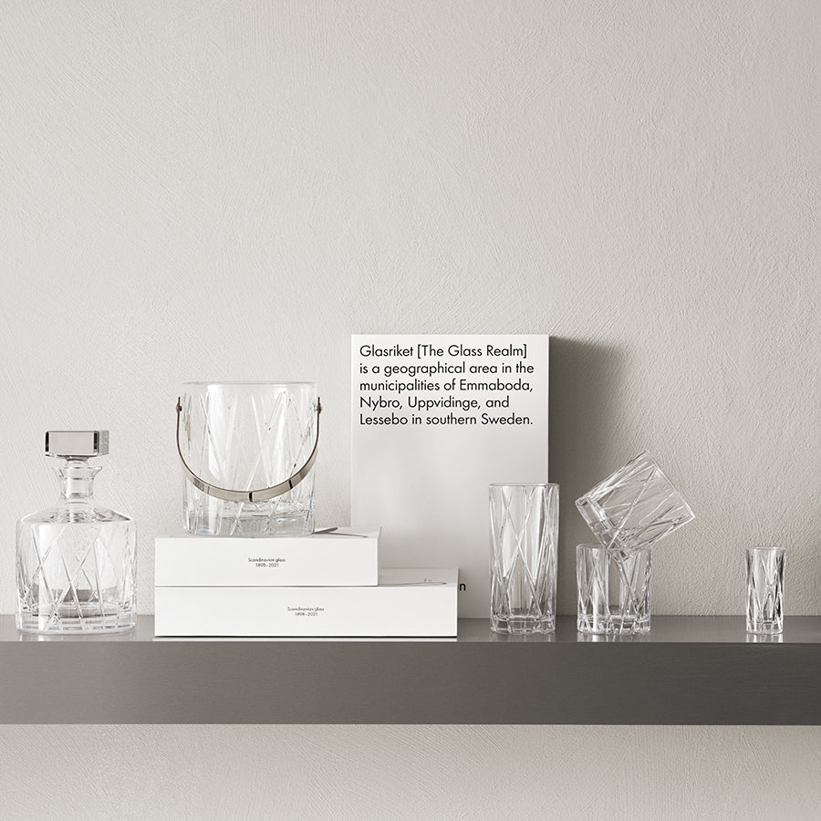 A shelf features glassware with a sign about Glasriket, the glass-making region in southern Sweden, showcasing an Orrefors: City Decanter 32oz, a vase, glasses, and neatly arranged boxes.