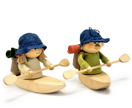 The Scandinavian Hiking Girl in Kayak is a wooden toy figure resembling a classic Scandinavian look, wearing a hat and carrying a rolled mat while paddling a wooden kayak with double paddles.