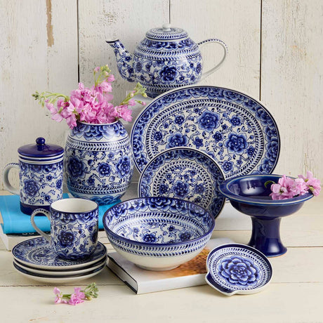 A collection of blue and white Bat Trang pottery, including dishes, cups, a teapot, and the Indigo Bloom Spoon Rest—some holding pink flowers—arranged on a white wooden surface.