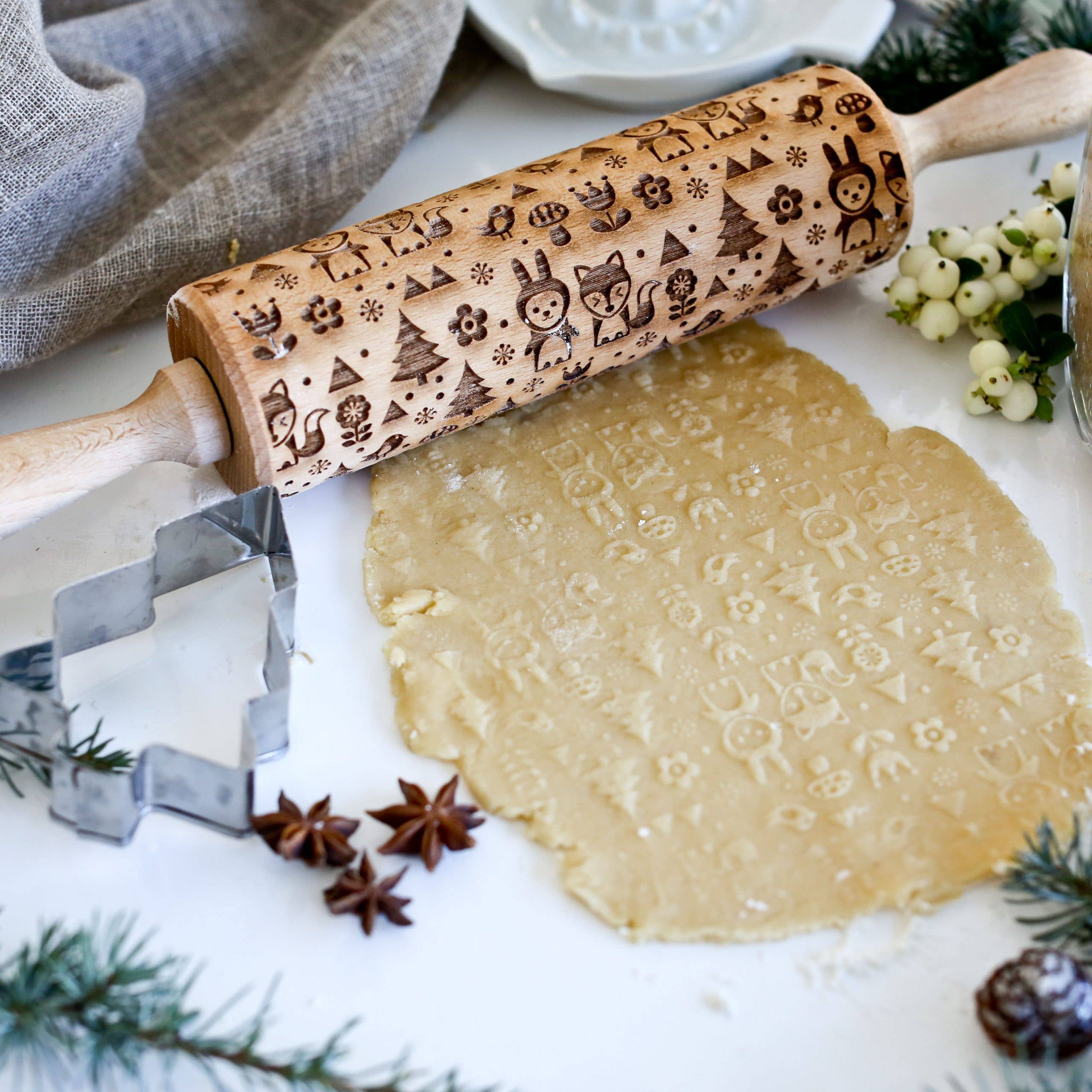 The Rolling Pin: Scandinavian 2 imprints a fox and rabbit pattern on cookie dough, styled with cookie cutters, spices, and greenery on a white surface.