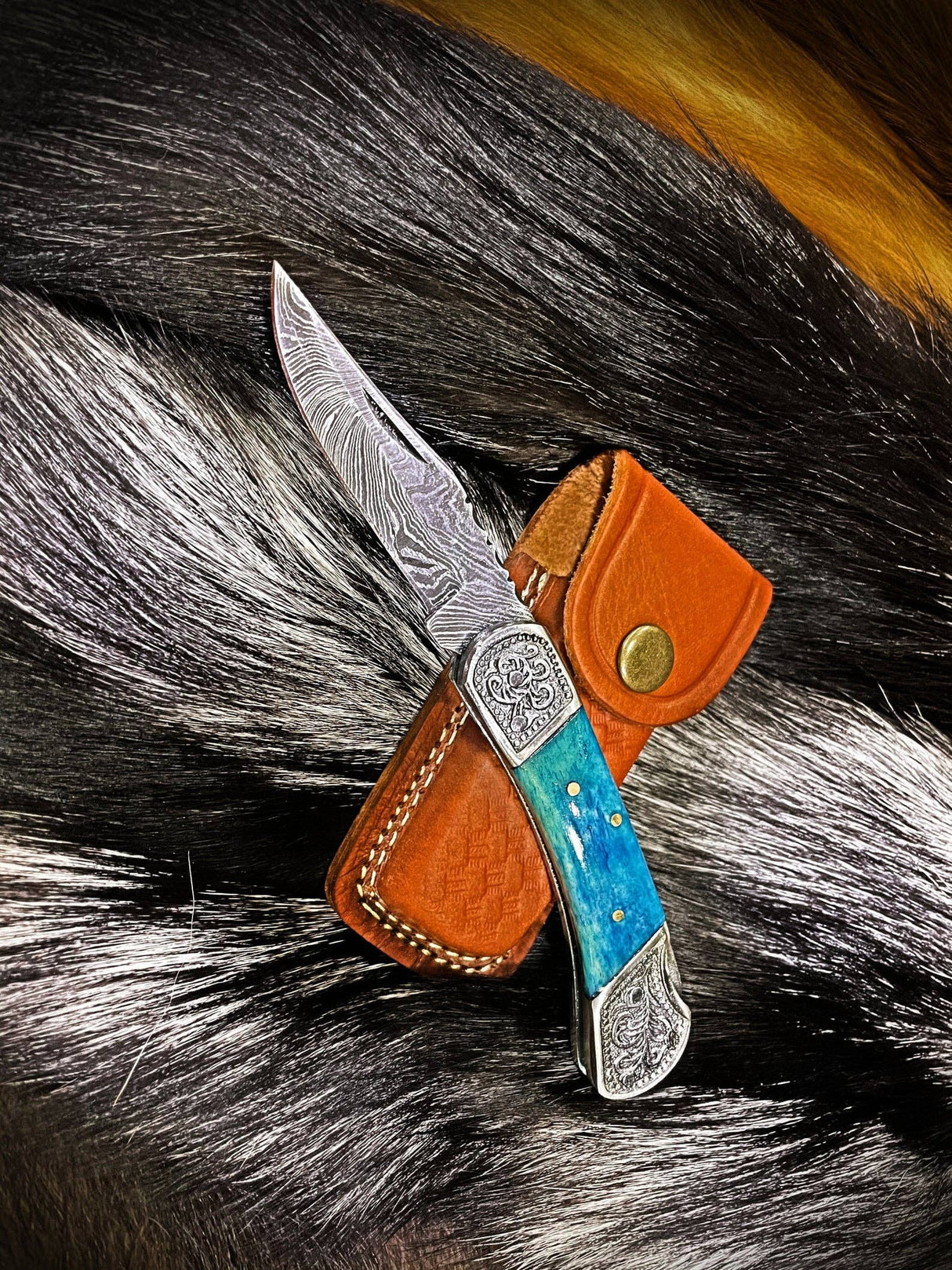 The Knife: Dyed Blue Bone 6.5 Damascus Steel features a patterned blade and blue bone handle, resting on a leather sheath, displayed atop animal fur.
