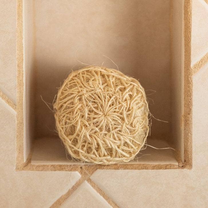The Scrub: Sisal Exfoliating Body Scrub, a round, natural twine ball designed for zero-waste beauty, rests in a recessed niche built into a tiled wall.