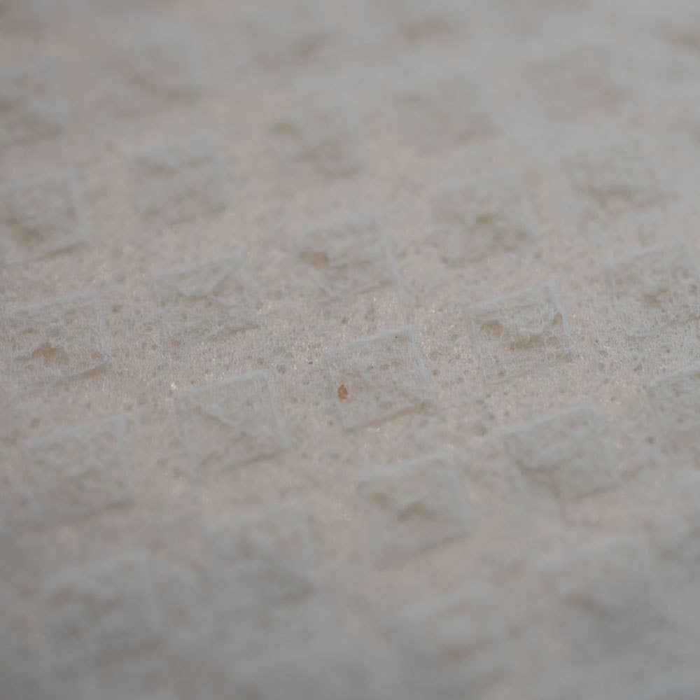 Close-up of the textured white surface of the SWEDEdishcloths Dish Cloth: Christmas Cardinal with Animals Swedish Dishcloth, featuring a repeating pattern of slightly raised diamond shapes. The lighting casts gentle shadows, emphasizing the three-dimensional texture characteristic of this reusable and sustainable cleaning cloth.