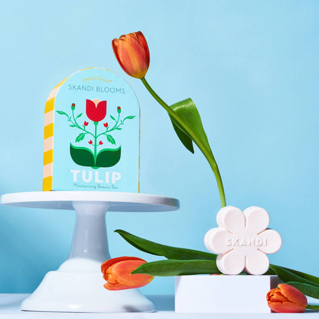 A flower-shaped Skandi Blooms Tulip Bar Soap by Swedish Dream stands beside its packaging, flanked by two orange tulips against a light blue background—an elegant, cruelty-free, and vegan beauty essential.