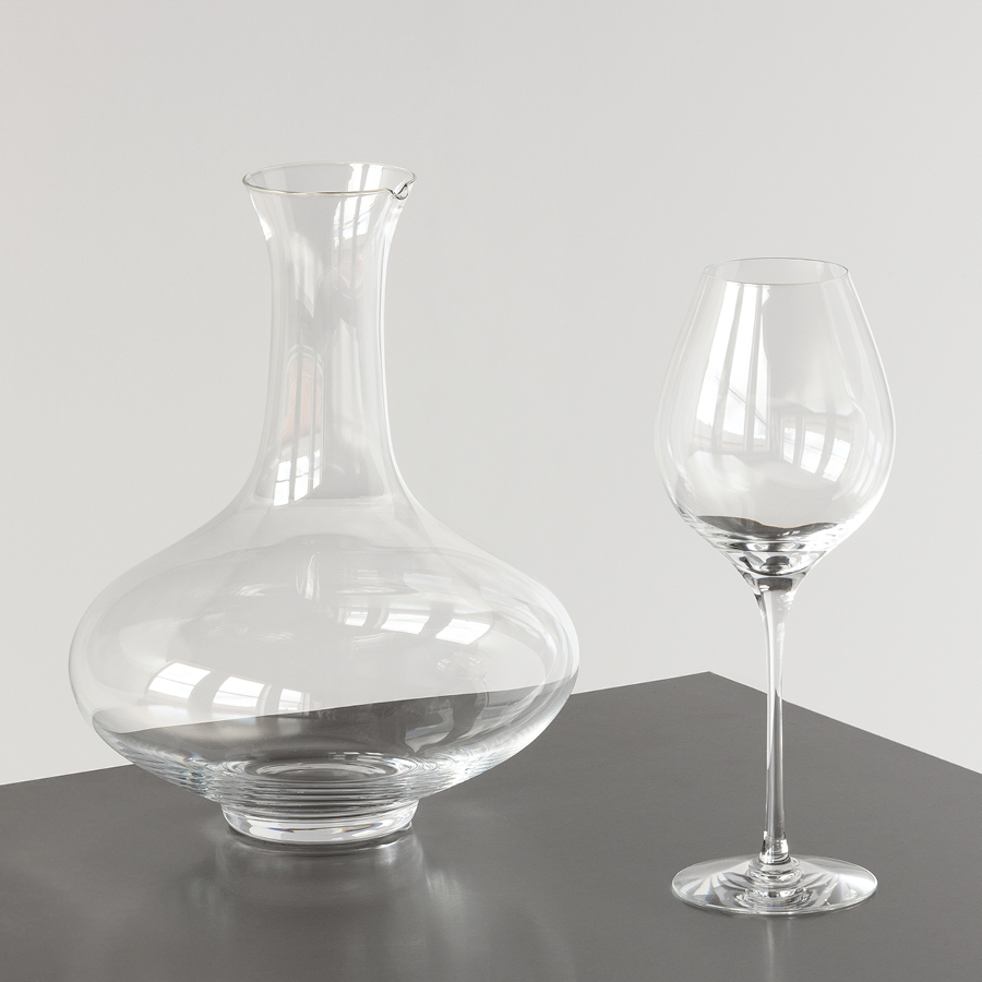 An empty Orrefors Difference Primeur Glass 21oz from the 2-pack is elegantly arranged beside a clear glass decanter on a grey surface against a light grey background.