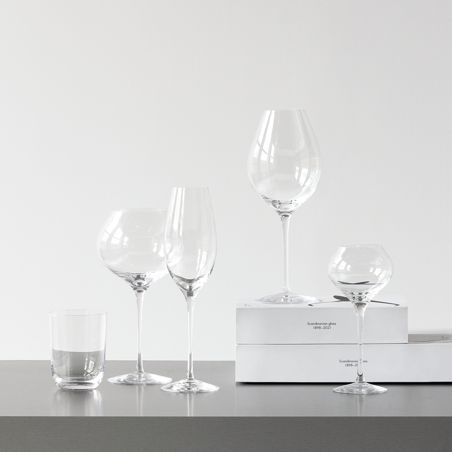 Six clear wine glasses, including the Orrefors Difference Sparkling Wine Glass 10oz, elegantly set on a gray surface. Two white boxes in the background suggest their exquisite Swedish mouth-blown origin.