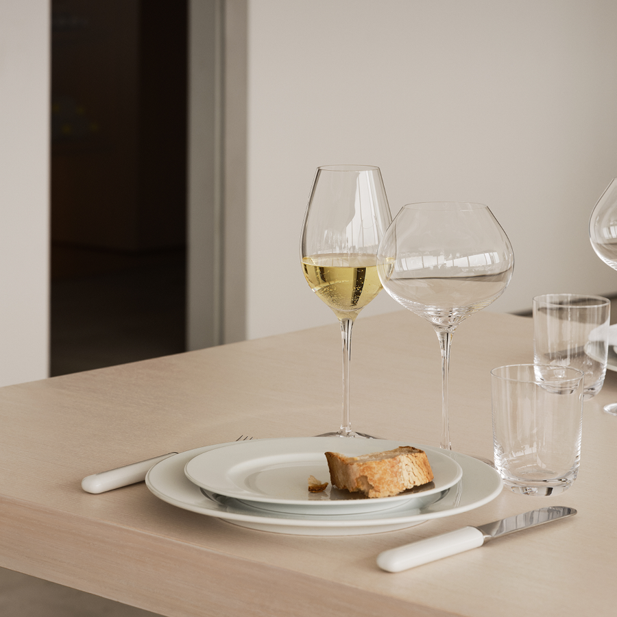 A dining table features a white plate, bread slice, and white-handled cutlery. Two Orrefors Difference Crisp Wine Glasses, 15oz each (2-Pack), designed by Erika Lagerbielke, hold crisp white wine with empty glasses nearby.