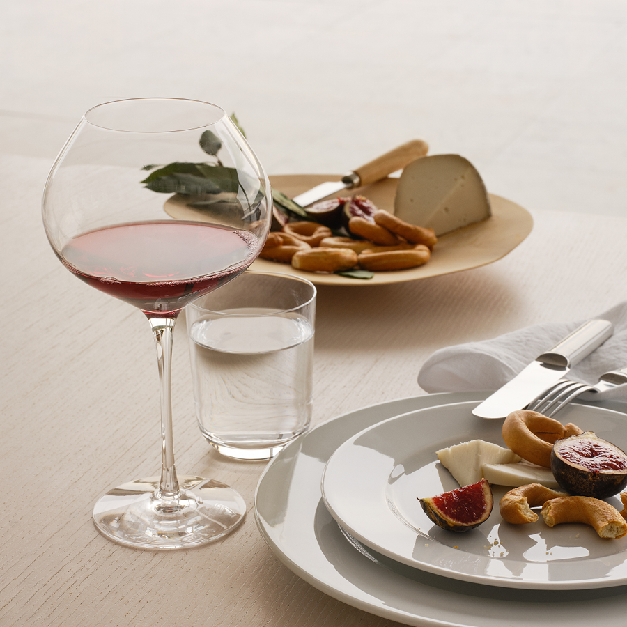 A glass of red wine, elegantly served in an Orrefors: Difference Mature Glass 22oz from the 2-Pack, sits next to a glass of water and a plate with cheese, figs, and crackers. In the background, another plate holds more cheese and breadsticks.