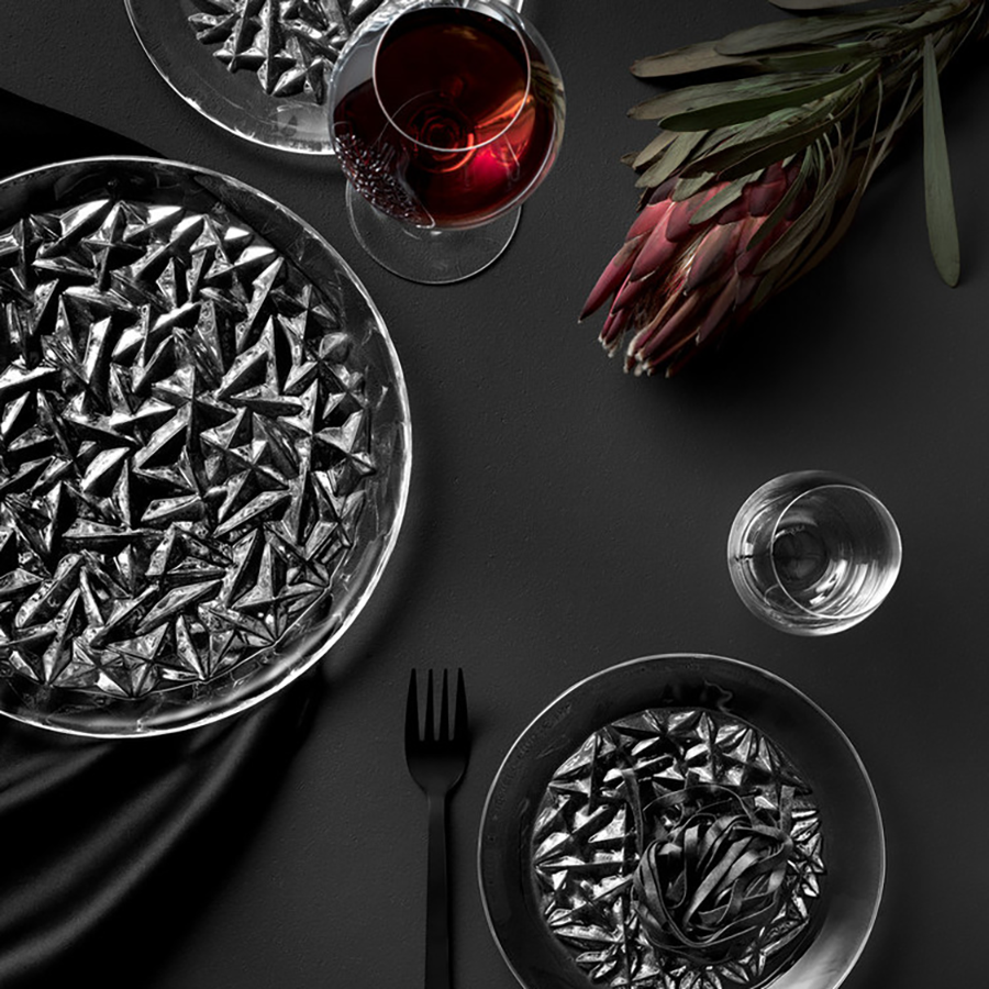 A table set with Orrefors: Carat Dinner Plates featuring geometric patterns, a crystal glass of red wine, and water on a dark surface. A floral arrangement in the top right suggests Lena Bergströms elegant design influence.