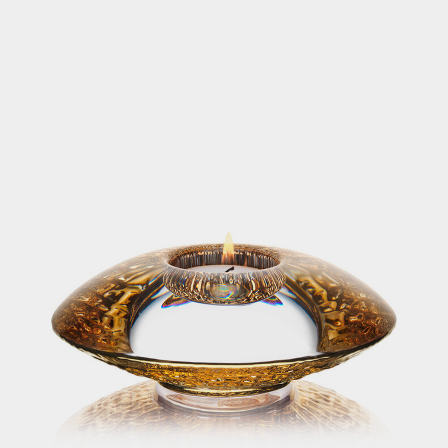 The Orrefors: Discus Gold Votive Candle Holder is a stunning glass piece featuring a round, flat, amber crystal design. Its reflective surface elevates any setting with elegance against a plain white background when lit.