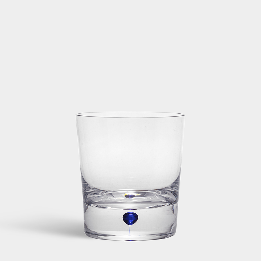 An Orrefors Intermezzo Blue Double Old Fashioned 13.5oz glass with a thick base and a small blue droplet inside is placed on a plain surface against a gray background.