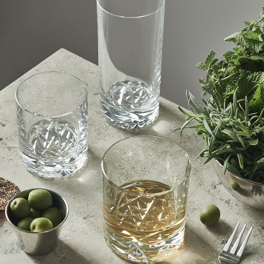 Three empty Orrefors: Carat Double Old Fashioned glasses and one filled with a light brown liquid rest elegantly on a textured countertop. A small cup of green olives and a bowl of fresh herbs nearby enhance the refined ambiance for any connoisseur of taste.