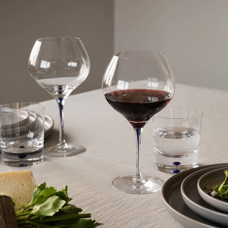 An Orrefors red wine glass adorns the table, alongside two empty Intermezzo Blue Double Old Fashioned glasses, water glasses, cheese plates, and fresh greens.