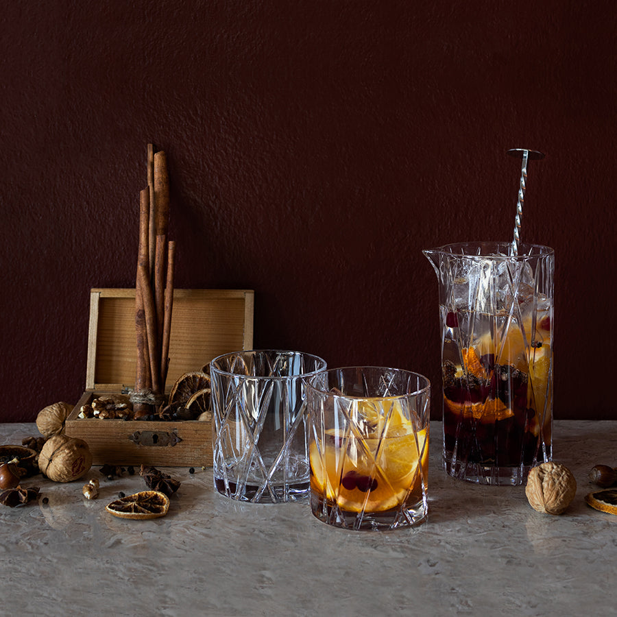 An Orrefors: City Double Old Fashioned Glass 11oz (2-Pack) holds a fruit-infused drink, resting on marble with cinnamon sticks, walnuts, and cloves against a dark brown backdrop.