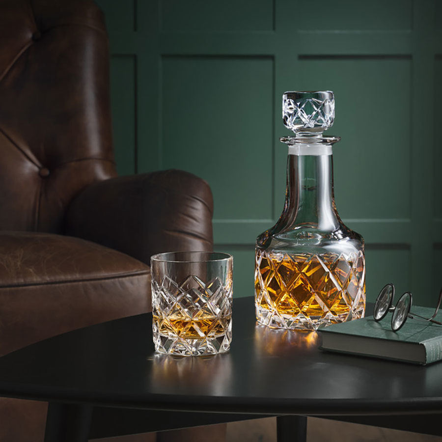 An Orrefors Sofiero Double Old Fashioned glass, filled with amber liquid, graces a table next to a leather chair, book, and glasses, showcasing timeless artistry.