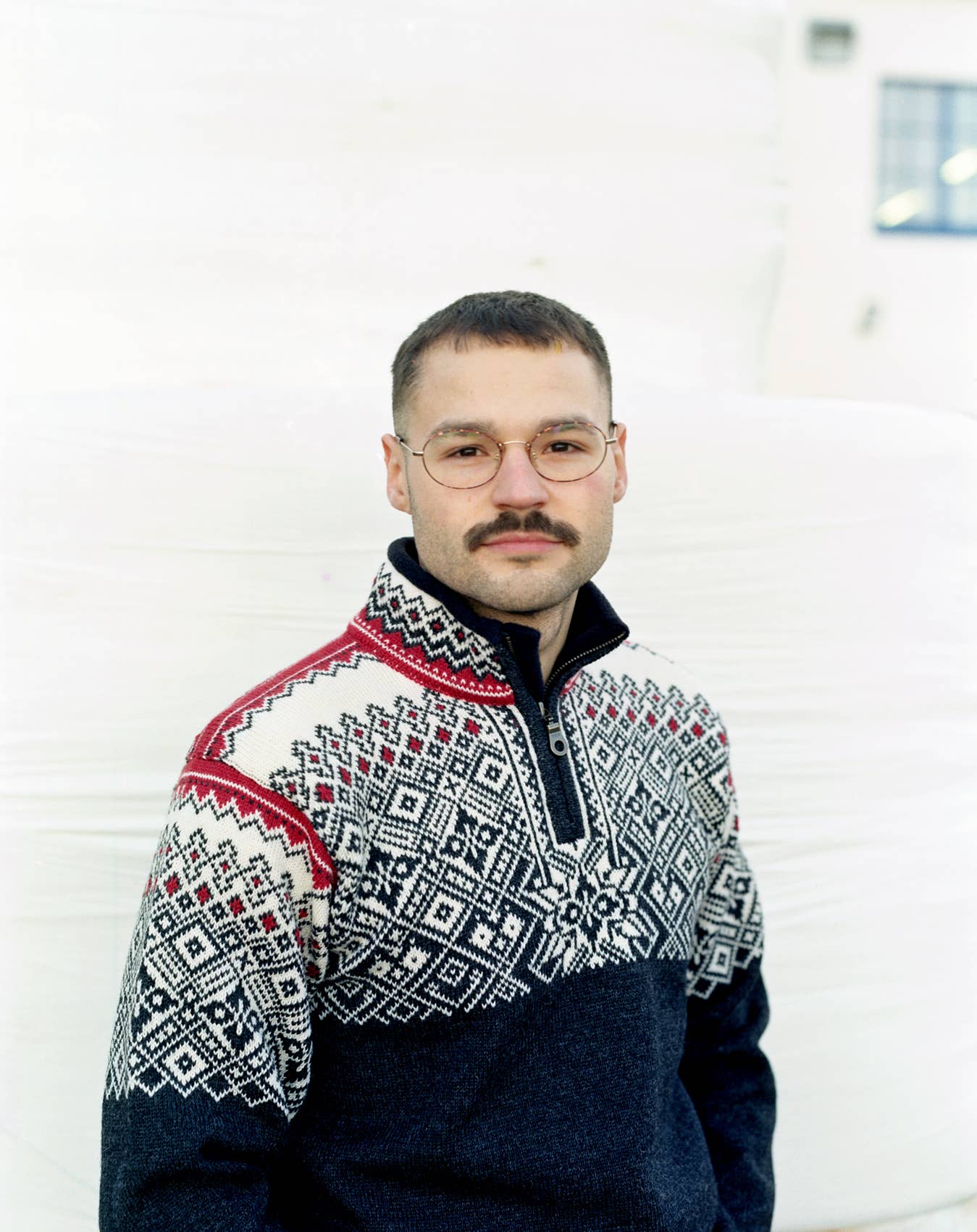 A man with a mustache and glasses is wearing the Narvik Ski Sweater in charcoal, patterned and made from 100% wool, standing in front of a white background.