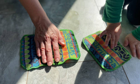 An adult and a child clean a tiled floor with Scrubbie: Skrubba XL Scrub Cloths, enjoying scratch-free scrubbing as sunlight casts shadows on their hands.