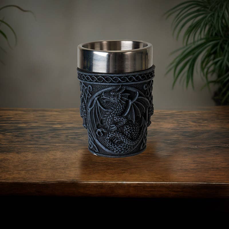 The Shot Glass: Medieval Dragon Shot Glass features a black ornate holder with a detailed dragon design and a stainless steel insert. It rests on wood, while blurred green plants appear in the background.