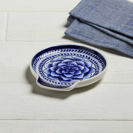 The Indigo Bloom Spoon Rest, a round ceramic dish with a blue floral Bat Trang-inspired pattern, rests on a white wooden surface beside folded blue cloth napkins.