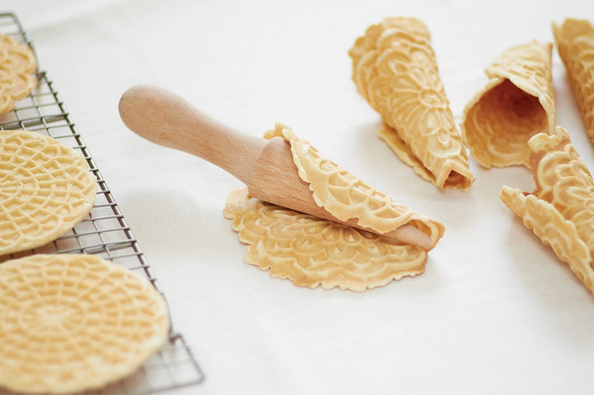 A Krumkake Cone Roller (Beechwood, 1.5" x 1.5" x 8.25") shapes a flat wafer into a crisp cone as finished pizzelle cones and extra wafers cool on the rack nearby.