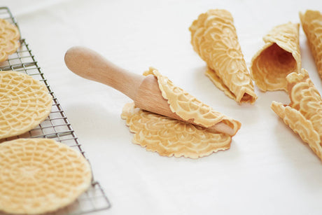 A Krumkake Cone Roller (Beechwood, 1.5" x 1.5" x 8.25") shapes a flat wafer into a crisp cone as finished pizzelle cones and extra wafers cool on the rack nearby.