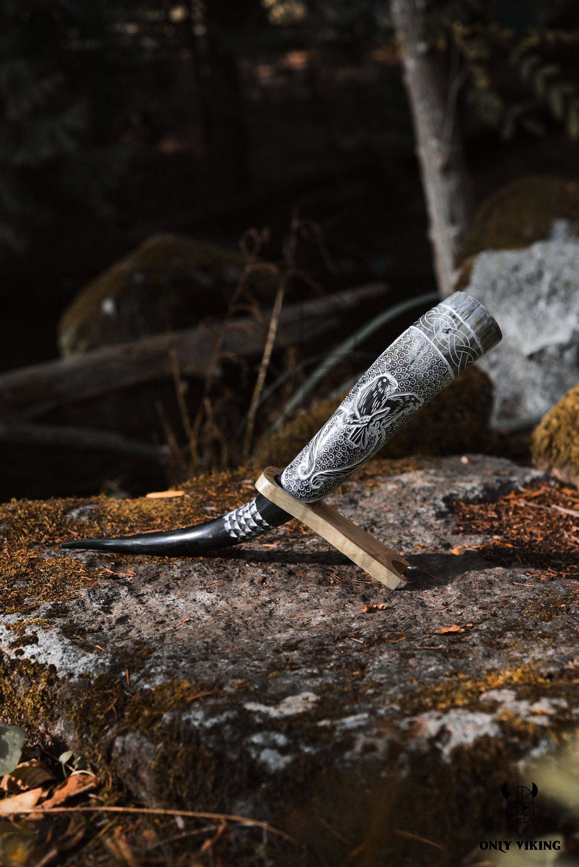 The Horn: Viking Drinking Horn w/ Metal Stand (14"-18") is elegantly displayed on a mossy rock in a forest setting, showcasing its intricate engravings reminiscent of raven motifs.