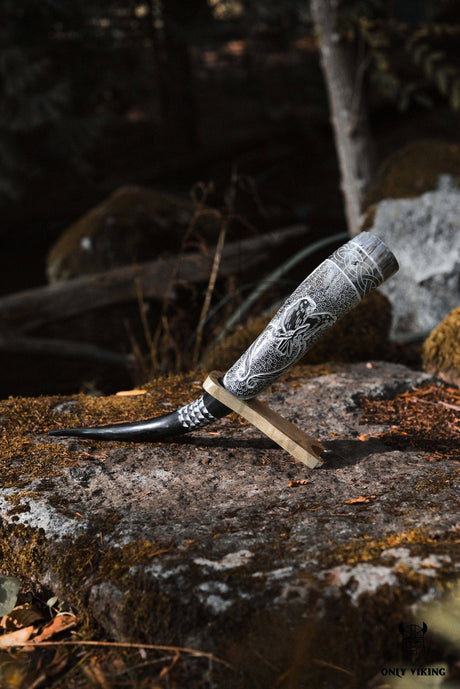 The Horn: Raven Viking Drinking Horn w/ Metal Stand (14"-18") features a carved raven motif and rests on its metal stand atop a mossy rock in a forest setting.