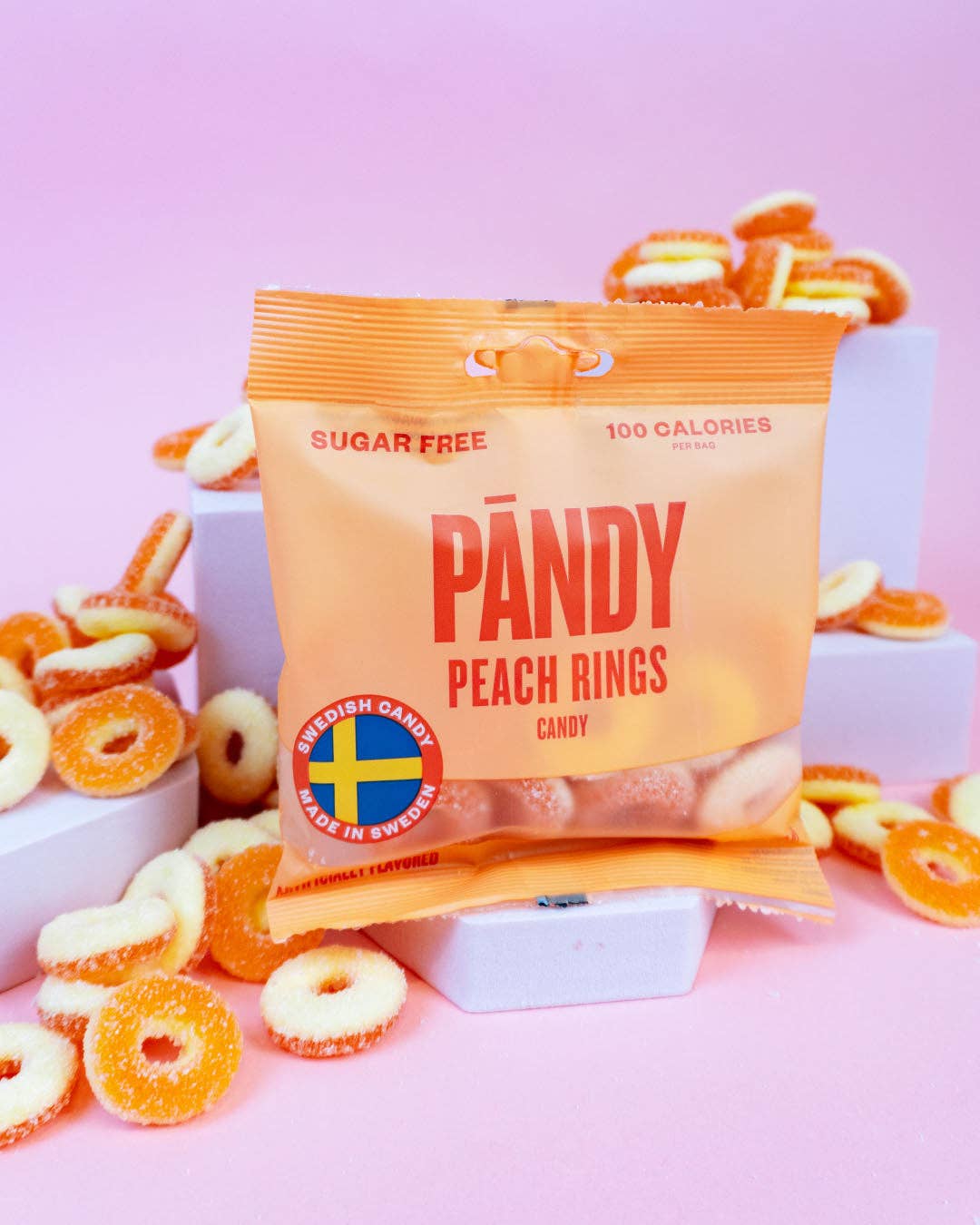 A bag of Pandy sugar free peach rings, weighing 1.8 ounces and imported from Sweden, is surrounded by loose candy on a pink background.