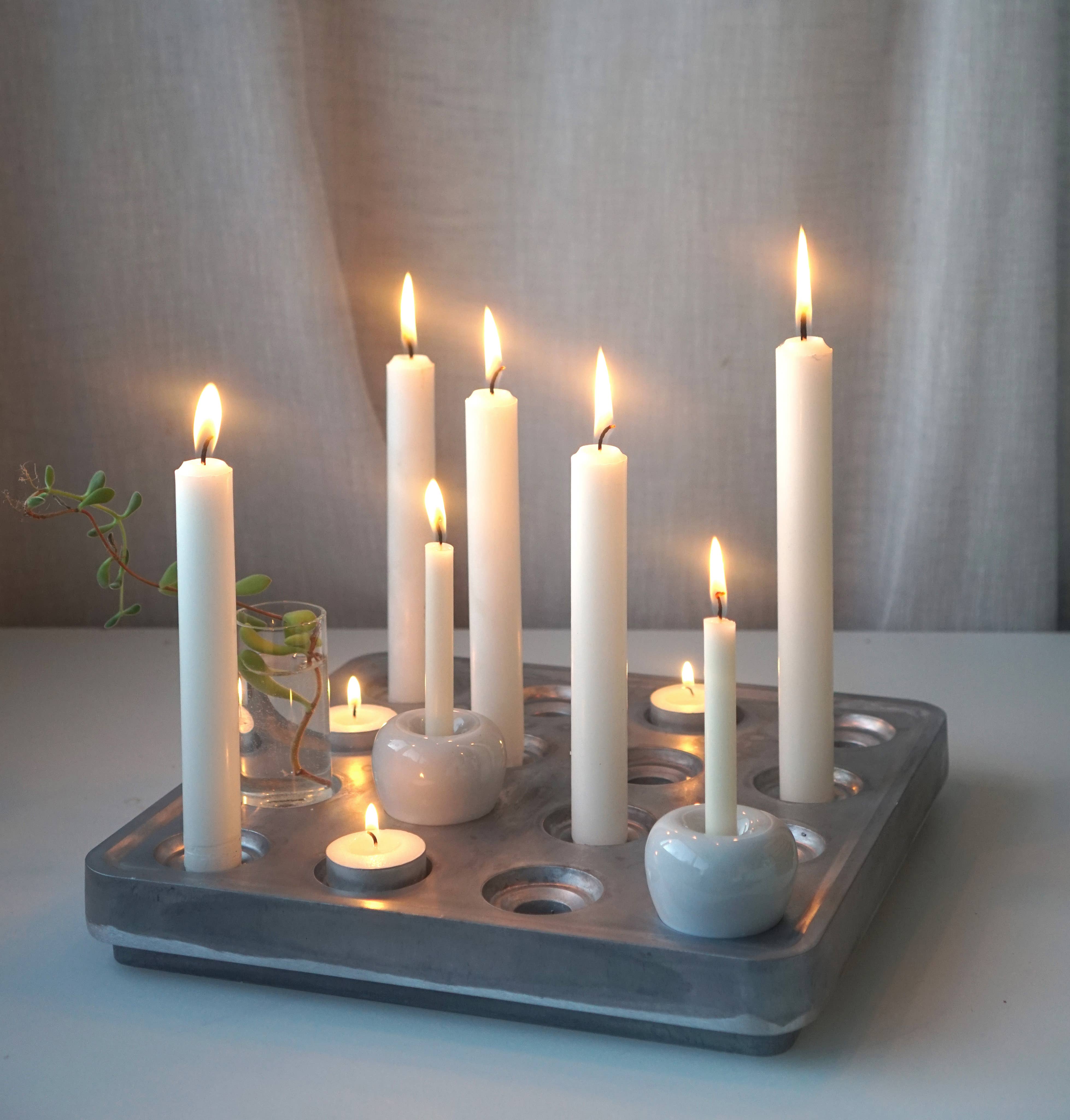 A pair of black mini apple candle holders are elegantly displayed on a sleek stoneware tray, accompanied by a small plant in a glass, against a light grey curtain backdrop.