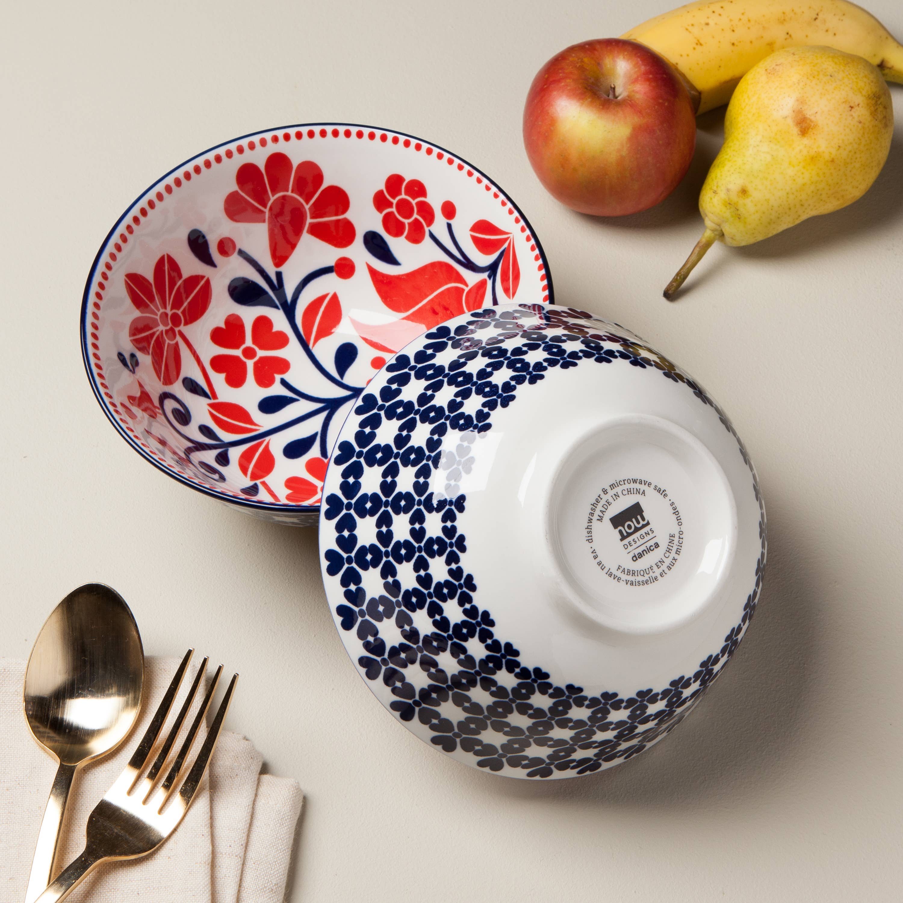 The Navy Bird Stamped Bowl, 6 inches in size, along with another decorative porcelain bowl featuring a floral pattern in blue, is displayed beside a pear, bananas, an apple, and gold-colored utensils on a napkin. These items exude cottage chic charm and add quaint elegance to any table setting.