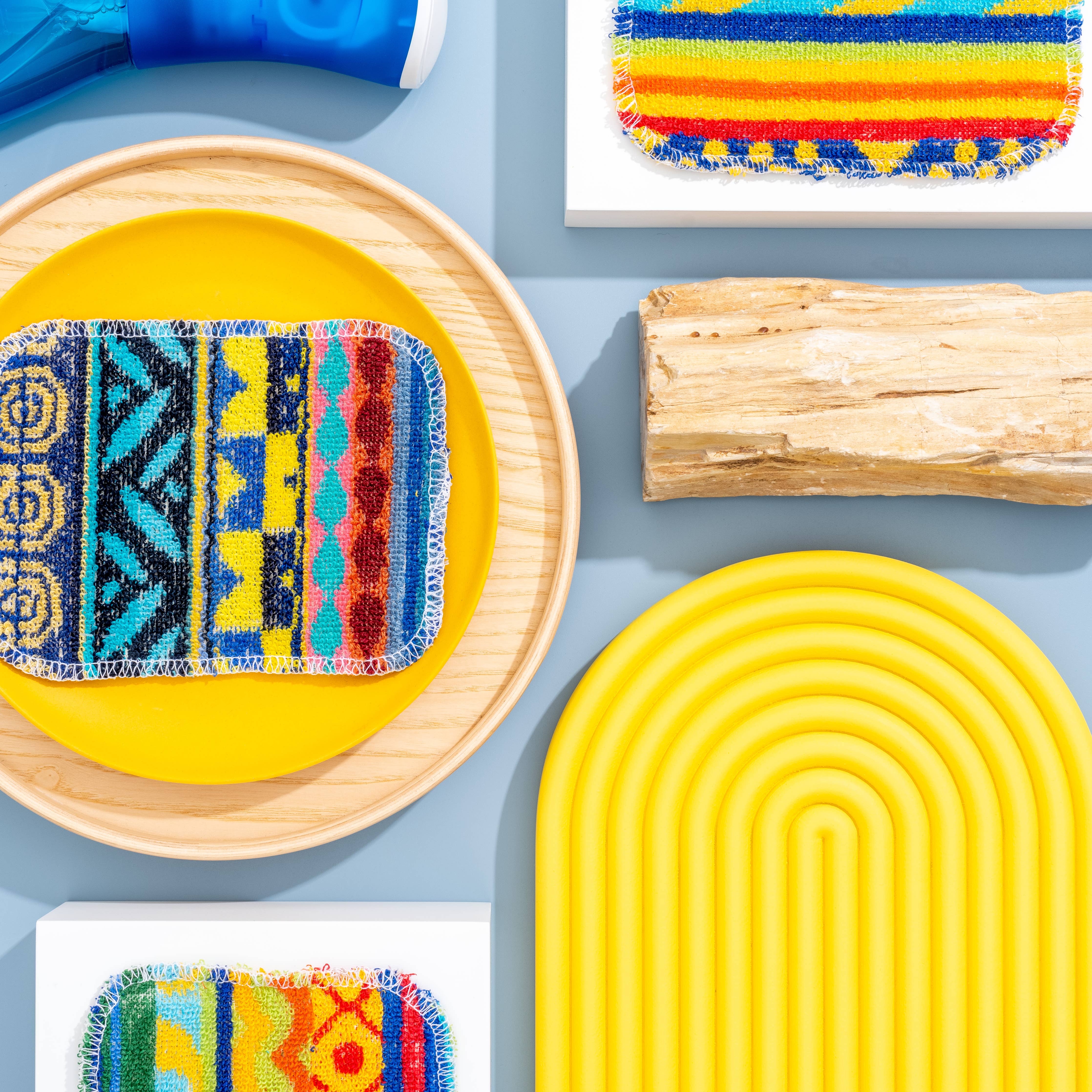 Euroscrubby Multi-Purpose Scrubber 3 Pack pads are artistically arranged on circular trays alongside a textured yellow mat, wooden log, and spray bottle against a light blue background. These eco-friendly scrubbers are ideal for effortless cleaning tasks.