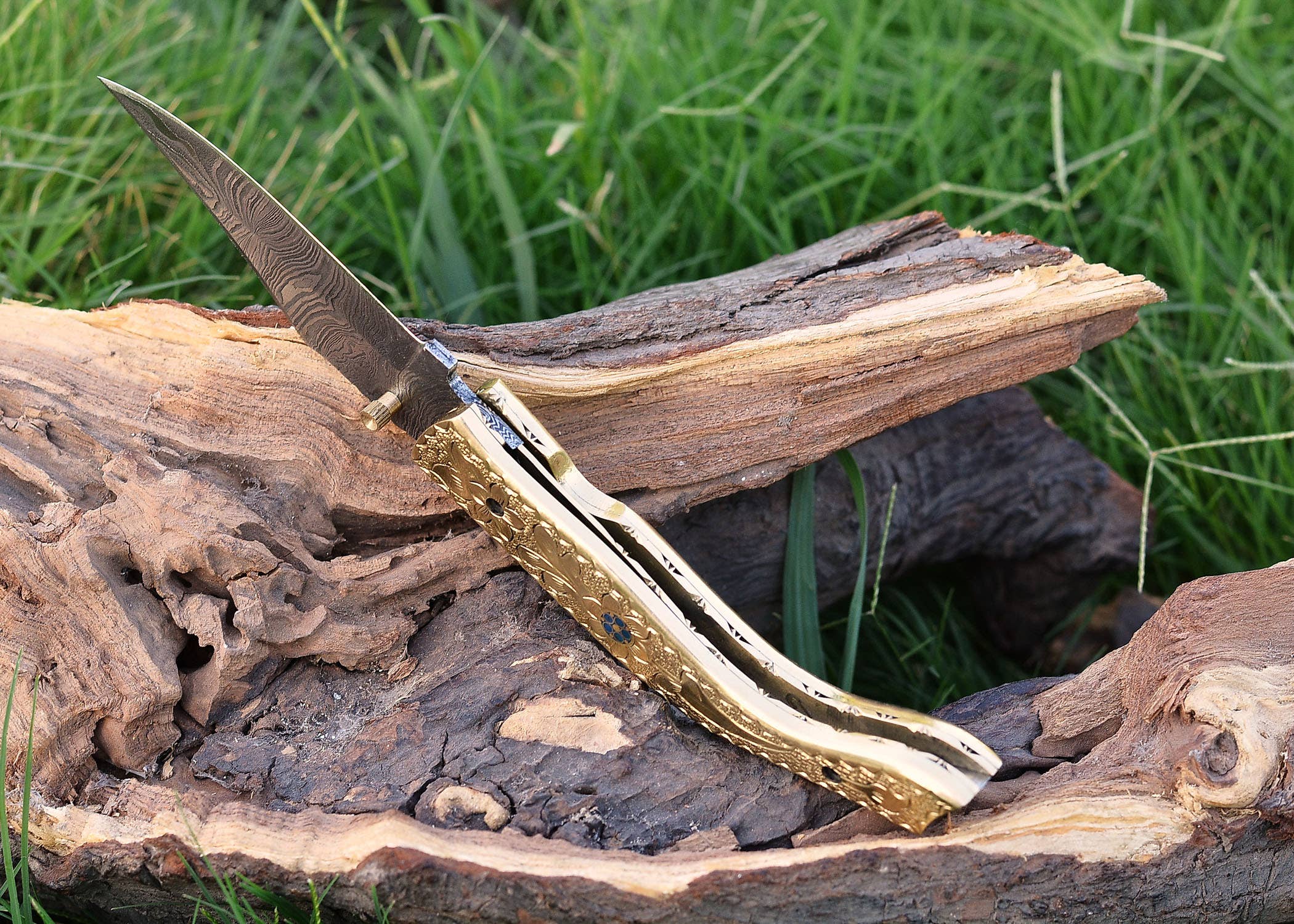 The Exquisite Golden Folding Knife, a luxury item with a stainless steel blade and an engraved golden handle, is elegantly displayed on weathered wood against a grassy background.