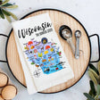 This vibrant Tea Towel, made in the USA, showcases a colorful map of Wisconsin with illustrations of notable landmarks and icons. Labeled The Badger State, it is displayed on a black tray alongside measuring spoons and eggs.