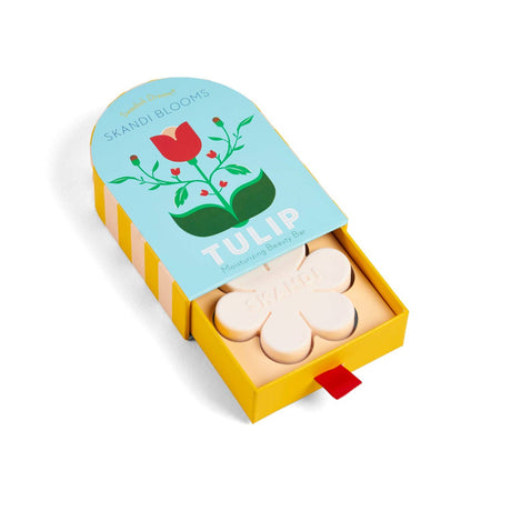 A yellow and blue box of Skandi Blooms Tulip Bar Soap by Swedish Dream is partially open, showing a flower-shaped soap inside. The packaging, with a red tulip illustration, highlights its gentle vegan formula for skincare.