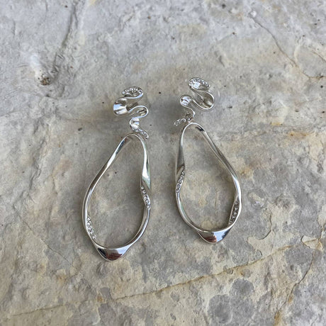 A pair of Blowing Ribbon Silver Dangle Earrings by A&C Oslo, featuring an irregular ribbon-like shape with small crystals, displayed on a textured stone surface.
