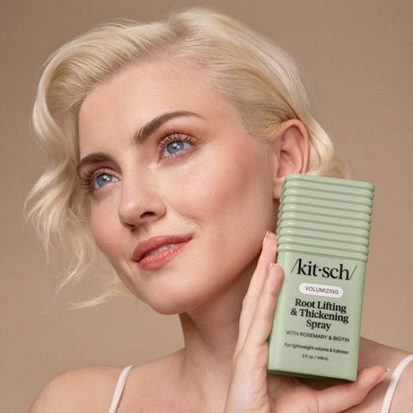 A person with short blond hair holds a green bottle of Kitsch Hair: Root Lifting & Thickening Spray 5 fl oz near their face, highlighting the volumizing spray against a neutral background.