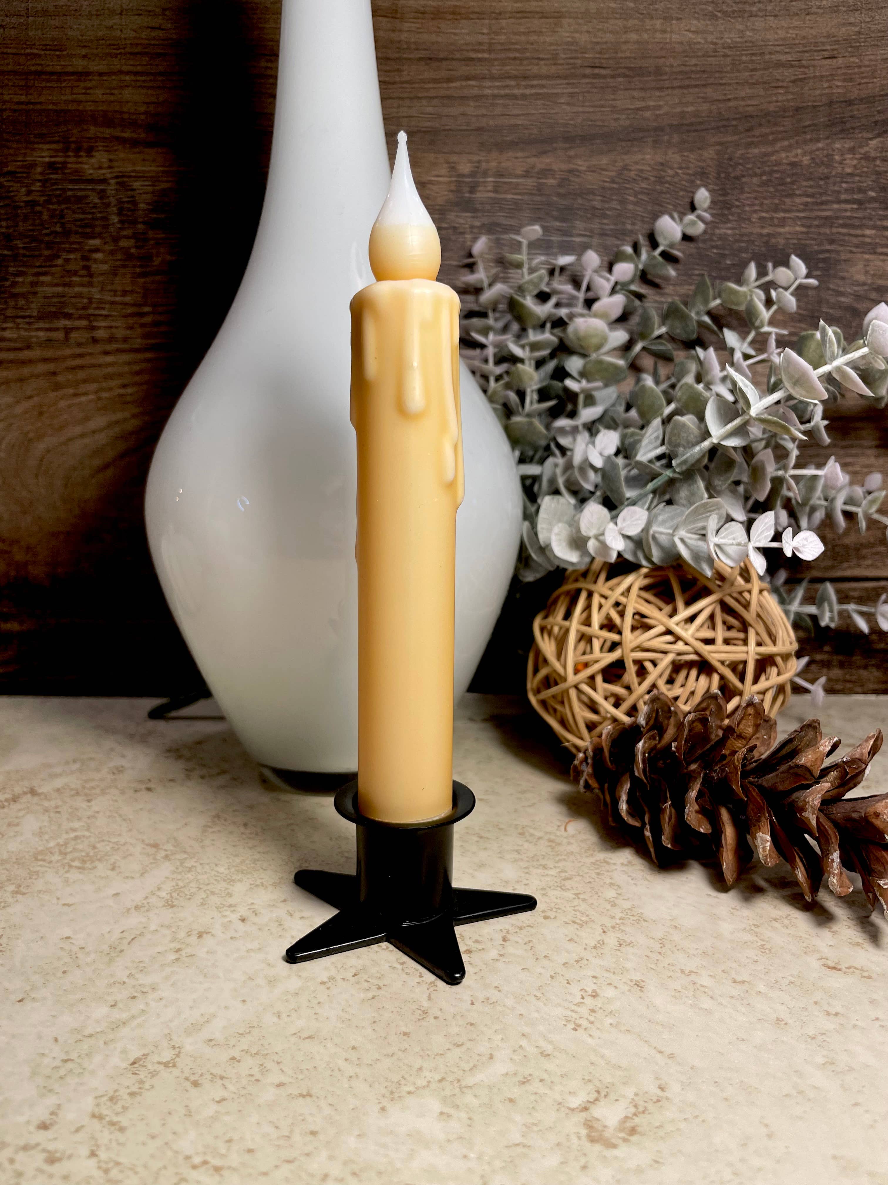 A tall, yellow candle with drip details is in the Star Wrought Iron Taper Holder. Placed on a light surface, it is paired with a white vase, greenery, and a pinecone for an elegant arrangement.