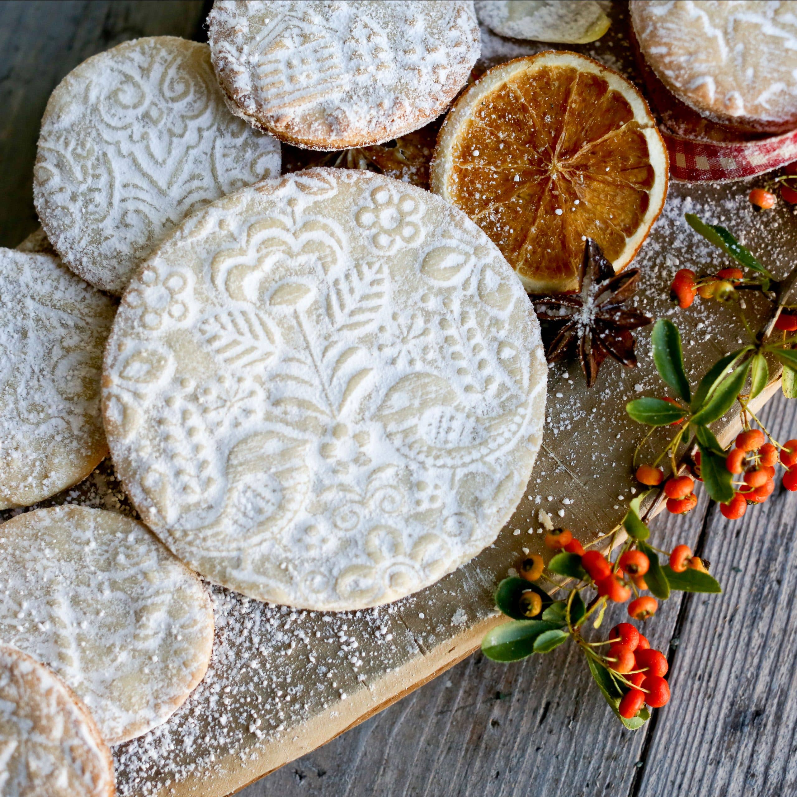 Patterned cookies made with the Scandinavian 4 - Birds and Trees Embossing Rolling Pin are dusted with powdered sugar, placed on a wooden board, and accompanied by dried orange slices, star anise, and small orange berries.