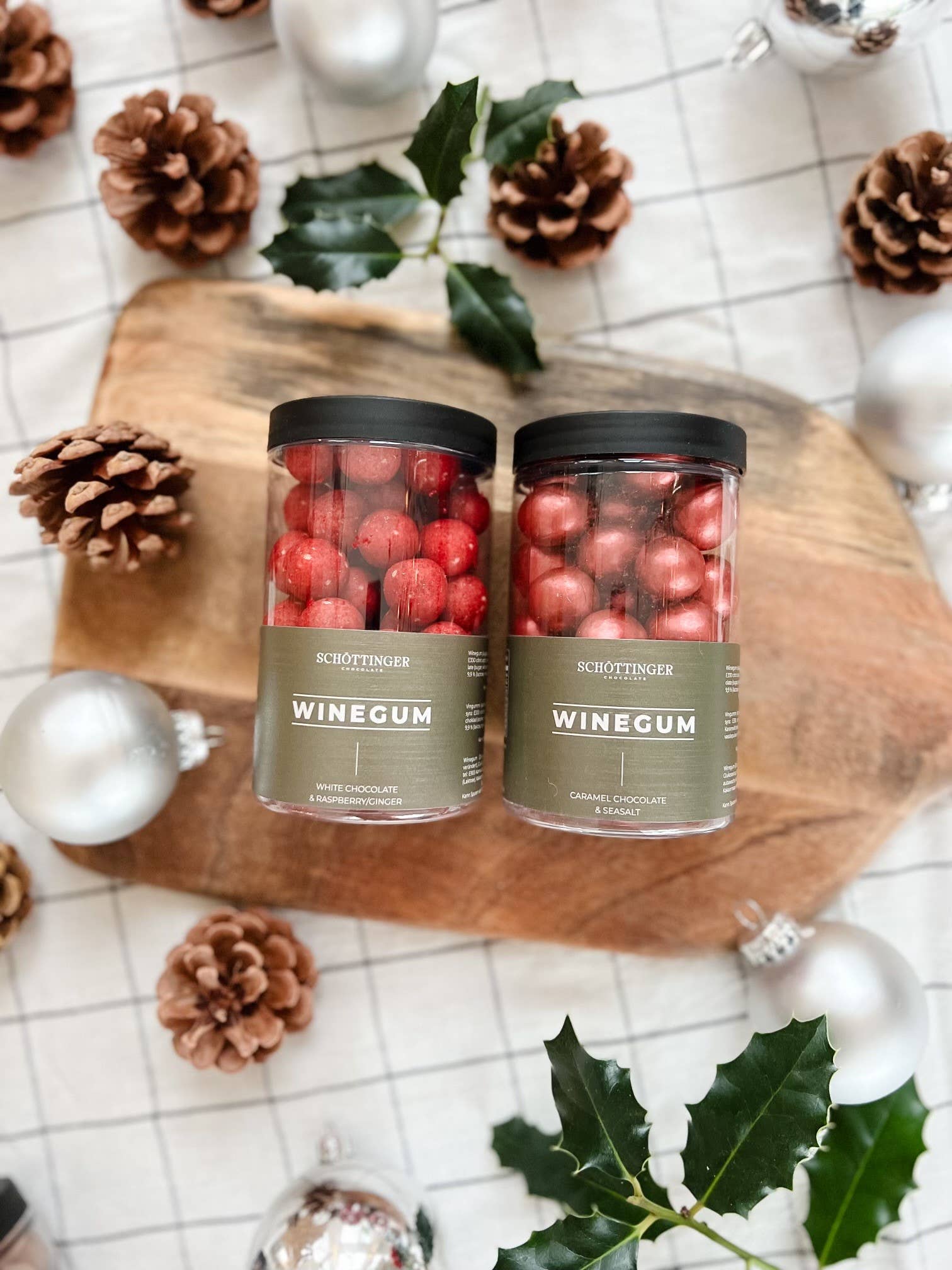 Two jars of Candy: Classic Winegum Seasalt & Caramel Chocolate (250 g) sit on a wooden board, surrounded by red amber pinecones, holly, and silver Christmas ornaments.