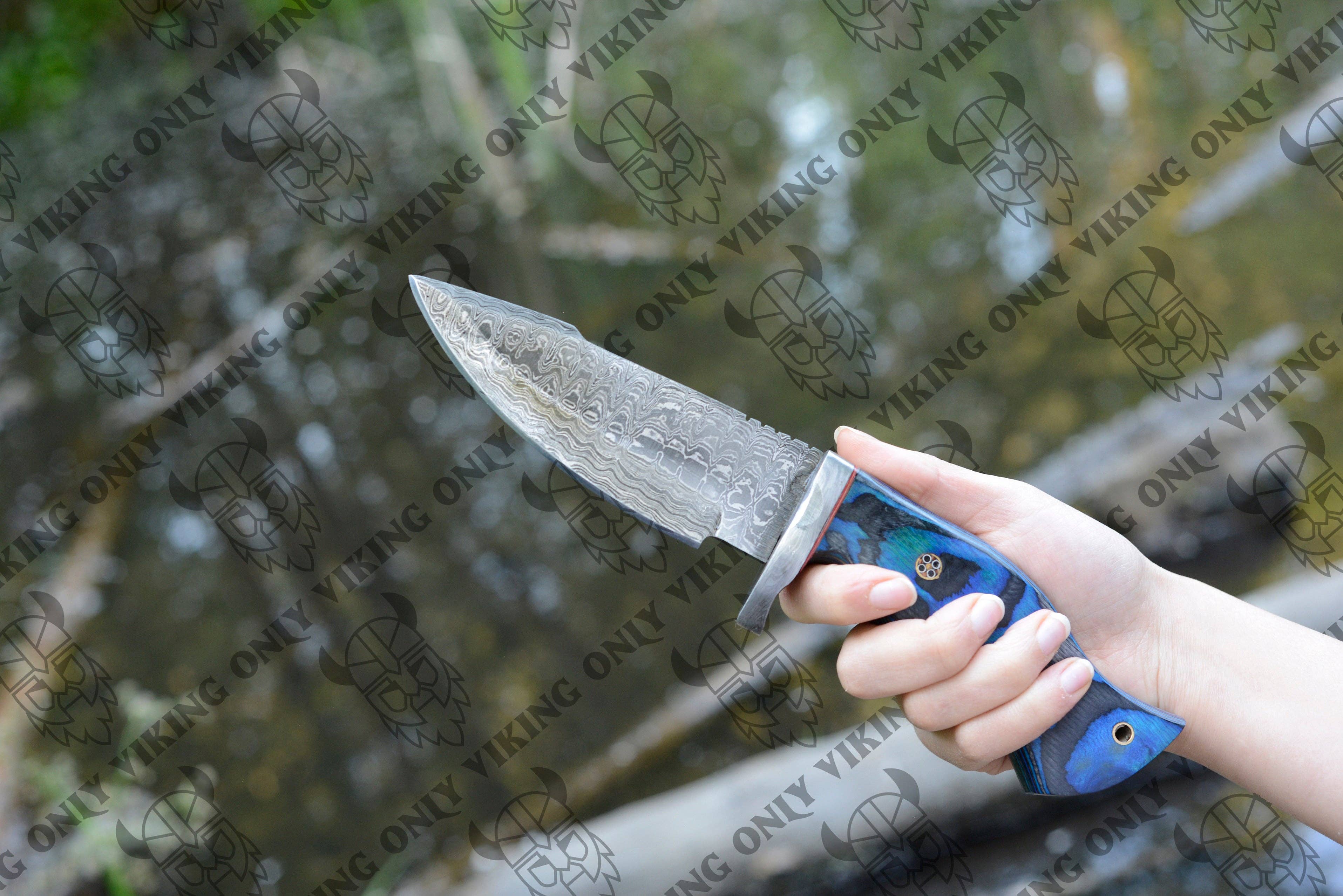 A hand holds the Knife: Premium Quality Damascus Utility Knife, featuring a patterned Damascus blade and a blue handle, outdoors near water.