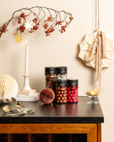 A tabletop features three jars of assorted chocolates, festive ornaments, a lit candle, and décor against a beige wall—highlighting Double Chocolate Danish Gourmet Licorice (Small, Limited Edition) for extra holiday indulgence.