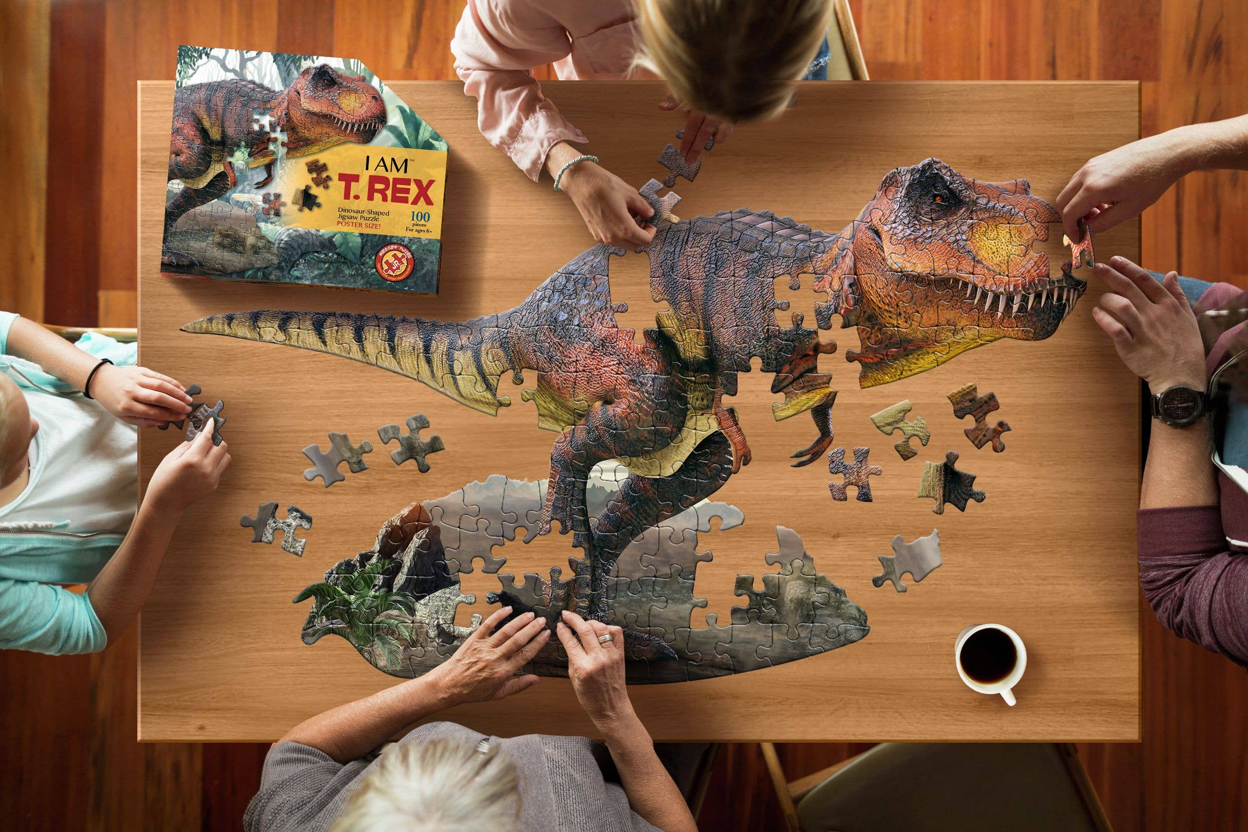 Three individuals are engrossed in assembling the I AM T. Rex 100 piece animal-shaped jigsaw puzzle, which depicts a T-Rex, on a wooden table. Nearby, a steaming cup of coffee sits beside the vibrant box labeled Puzzle: I AM T. Rex 100 piece, adding to the scene's charm and blending creativity with educational fun facts about these mighty creatures.