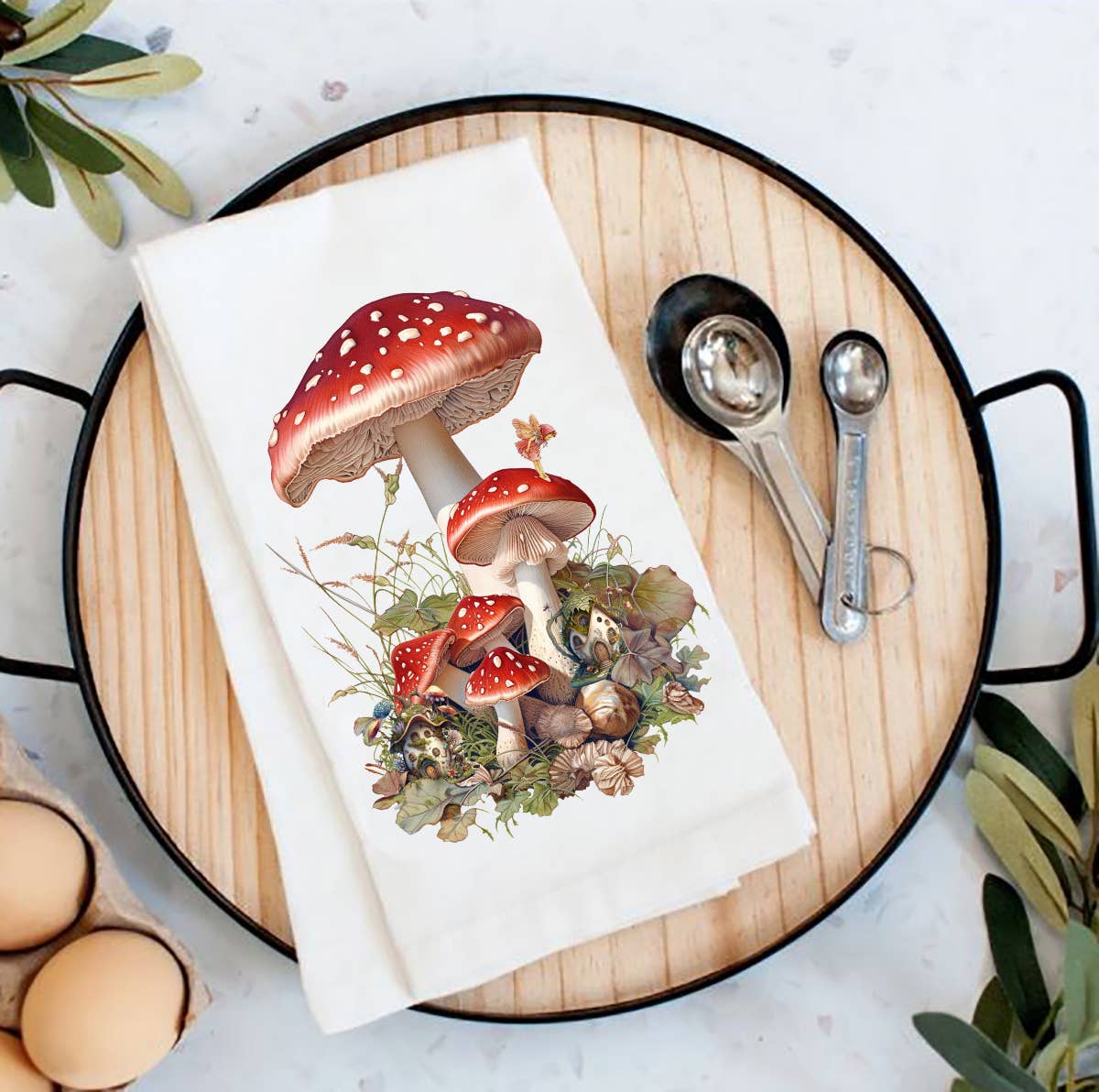 A round wooden tray with a charming Red & White Mushrooms with Fairies flour sack tea towel makes a perfect housewarming gift. Measuring spoons, eggs, and leaves are artfully arranged around it, creating a cozy and inviting kitchen atmosphere.