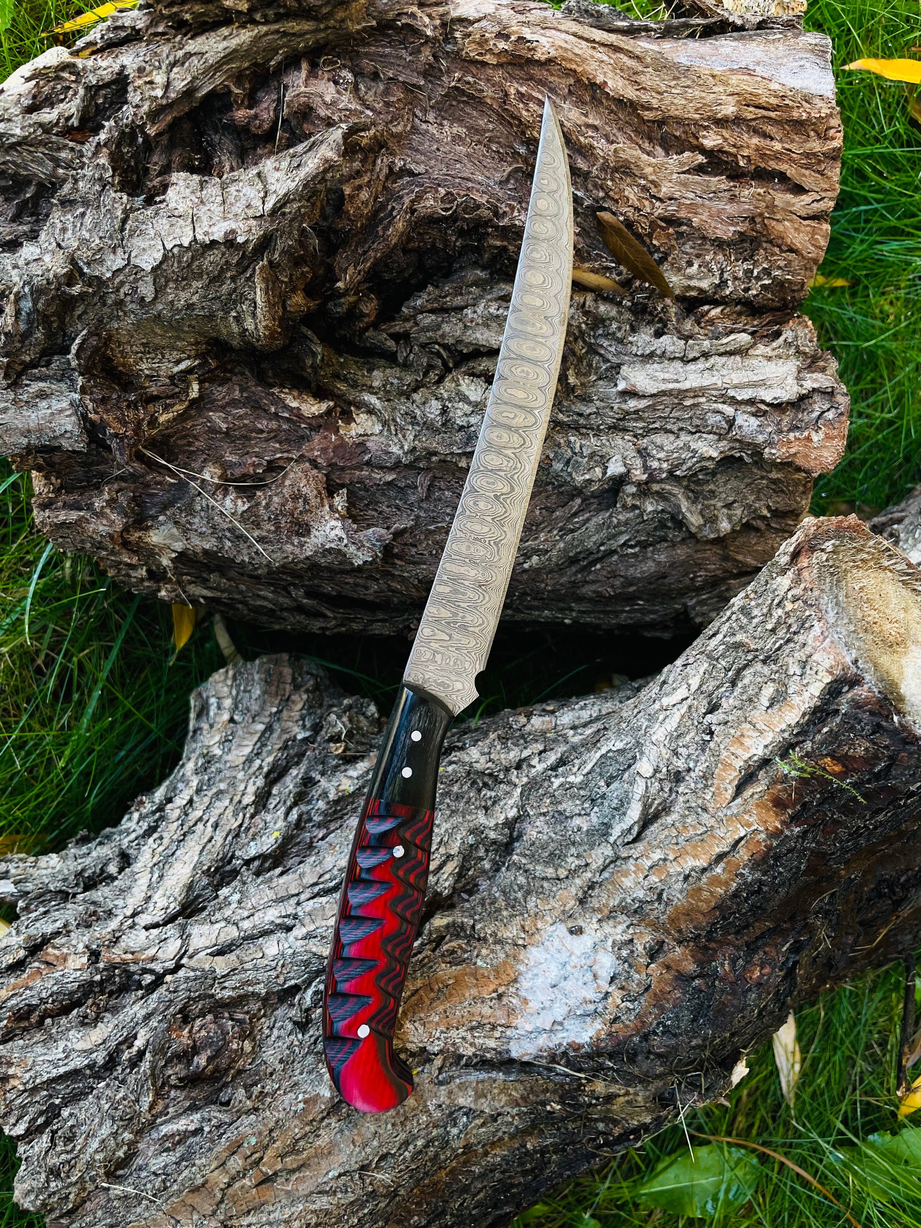 A Knife: Damascus Steel Custom Flay Knife with carved grips, featuring a patterned blade and red-black handle, is placed on rough tree bark outdoors on grass.