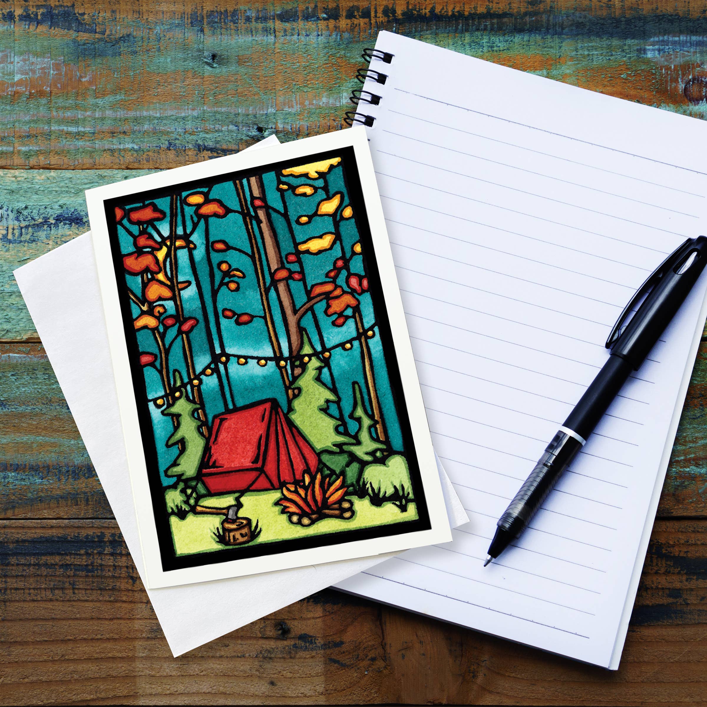 Notebook and pen beside a vibrant Sarah Angst Art greeting card titled "Evening at Camp Tent," featuring a red tent, trees, and string lights in a whimsical forest scene.