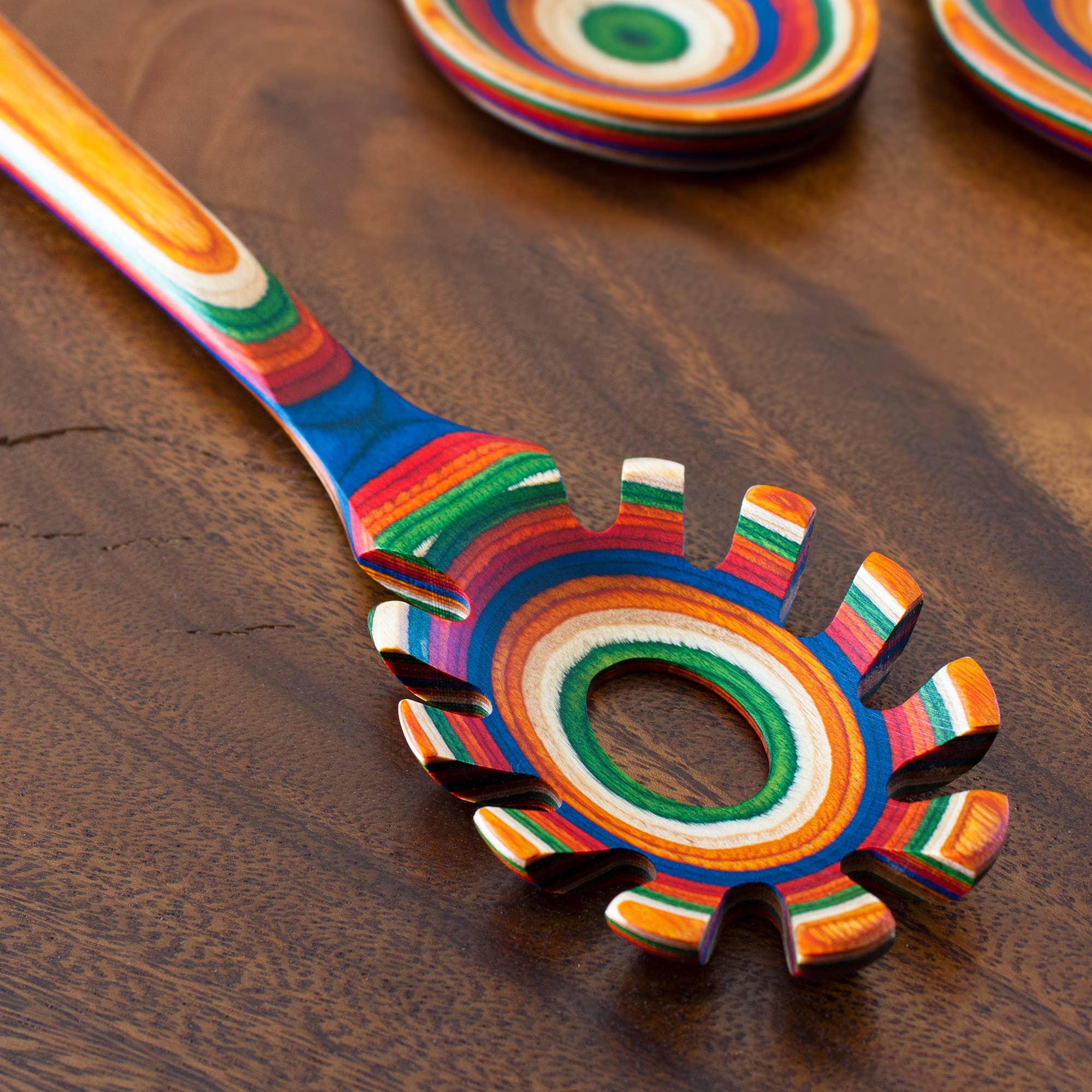 A vibrant Utensil: Marrakesh Collection Spaghetti Server, adorned with colorful stripes and featuring a central hole, rests gracefully on a wooden surface, showcasing the practicality of this non-stick kitchen tool.