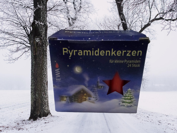 A box of Candles: Christmas Pyramid Candles features a winter scene design with a snowy landscape and a red star-shaped window, set against a background of a snowy path lined with trees. This 24-pack includes red candles that perfectly embody the festive spirit.