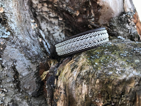 The Traditional Sami Bracelet #5B, crafted from black reindeer leather with pewter and silver thread, features intricate patterns and rests on the rough bark of a tree.