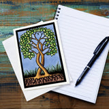 A Card: Together Forever Tree by Sarah Angst 5x7 rests on a blank envelope next to an open lined notebook and a black pen on a wooden surface. Made in America.